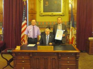 Governor Terry Branstad presents Regis Zweigart, ACS, ALB, Area 34 Director for District 19, and Paul Wood, DTM, Immediate Past District 19 Director, with the signed proclamation that October is Toastmasters Month